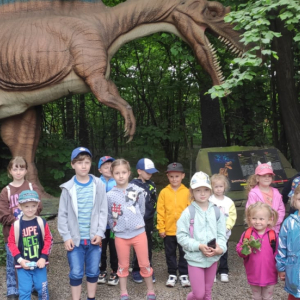 Przedszkolaki z Rudy Rysia w Parku Dinozatorland. Dzieci stoją w grupie. W tle makieta dinozaura. Kliknięcie na zdjęcie spowoduje jego powiększenie do rozmiaru oryginalnego.