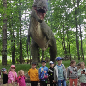 Przedszkolaki z Rudy Rysia w Parku Dinozatorland. Dzieci stoją w grupie. W tle makieta dinozaura. Kliknięcie na zdjęcie spowoduje jego powiększenie do rozmiaru oryginalnego.