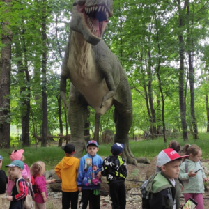 Przedszkolaki z Rudy Rysia w Parku Dinozatorland. Dzieci stoją w grupie. W tle makieta dinozaura. Kliknięcie na zdjęcie spowoduje jego powiększenie do rozmiaru oryginalnego.