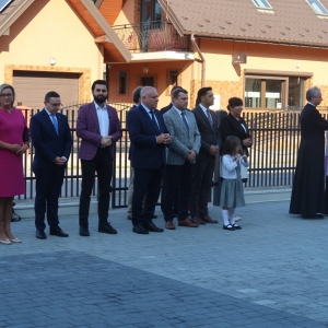 Przedstawiciele rządu RP, władz wojewódzkich, samorządowych, Dyrekcji, nauczyciele, fotografowie stoją przed głównym wejściem do budynku Zespołu Szkół w Szczurowej. Goście ubrani w galowe stroje. Obok ksiądz probosz Parafii Szczurowa wraz z proboszczem Parafii Rudy Rysie. Ks proboszcz czyta modlitwę dotyczą poświęcenia budnyku Zespołu Szkół w Szczurowej. Kliknięcie na zdjęcie spowoduje jego powiększenie do rozmiaru oryginalnego