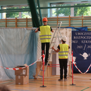 Występ uczniów Zespołu Szkół w Szczurowej. Dzieci przebrane za robotników budowlanych. Ubrani w kaski i żółte odblaskowe kaski. W tle dekoracja.   Kliknięcie na zdjęcie spowoduje jego powiększenie do rozmiaru oryginalnego