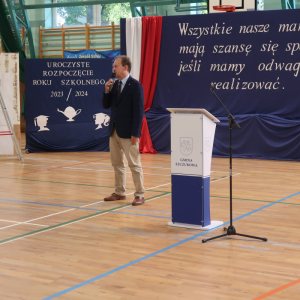 Przemówienie pana wiceministra edukacji Włodzimierza Bernackiego. W tle biało-niebieska dekoracja z biało-czerwoną flagą Polski. Przedstwaiciele rządu RP, władz wojewódzkich, samorządowych, Dyrekcji, nauczyciele, fotografowie stoją przed głównym wejściem do budynku Zespołu Szkół w Szczurowej. Goście ubrani w galowe stroje. Obok pani Dyrektor Joanna Ozorka-Krawczyk.  Kliknięcie na zdjęcie spowoduje jego powiększenie do rozmiaru oryginalnego