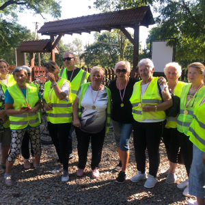 Seniorzy z klubu Seniora w Szczurowej stoją w szeregu na terenie otaczającego dworek w Strzelcach Wielkich. Ubrani są w żółte odblaskowe kamizelki i sportowe ubrania. Kliknięcie na zdjęcie spowoduje jego powiększenie do rozmiaru oryginalnego.