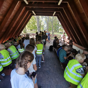 Uczestnicy rajdu rowerowego "Pokolenie ma moc" siedzą pod wiatą i spożywają kiełbaski. Seniorzy, uczniowie i nauczyciele ubrani są w kamizelki odblaskowe i letnie ubrania. Kliknięcie na zdjęcie spowoduje jego powiększenie do rozmiaru oryginalnego.