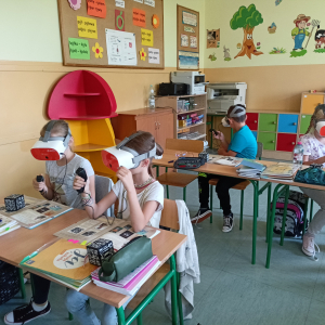 Lekcja w kl. 3b z wykorzystaniem okularów VR. Dzieci siedzą w sali lekcyjnej, na głowie mają założone okulary Vr. Oglądają materiał obrazkowy związany z II wojną światową. Poruszają się w wirtualnych okopach wojskowych za pomocja joysticków. Kliknięcie na zdjęcie spowoduje jego powiększenie do rozmiaru oryginalnego.