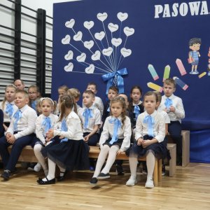 Uczniowie kl. I a  na pasowaniu. Dzieci siedzą na ławeczkach, ubrane w biało-czarne lub granatowe stroje. Mają też zawiązane granatowe kokardy na szyi. W tle granatowo biała dekoracja z białym napisem i ozdobnymi elementami. Kliknięcie na zdjęcie spowoduje jego powiększenie do rozmiaru oryginalnego.