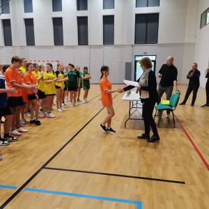 Wręczanie dyplomów i nagród w hali sportowej w Dębnie. Uczestnicy ubrani w kolorowe stroje sportowe stoją w szeregu. Reprezentantka drużyny dziewcząt z Zespołu w Szczurowej odbiera dyplom. Wręcza ją Dyrektor szkoły w Dębnie. Kliknięcie na zdjęcie spowoduje jego powiększenie do rozmiaru oryginalnego.