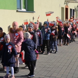 Uczniowie Zespołu Szkół w Szczurowej idą w parach na plac przed głównym wejściem do szkoły. Dyrekcja, nauczyciele i księża ubrani w jesienne ubrania idą wzdłuż budynku szkolnego. Uczestnicy akcji trzymają w rękach biało-czerwone chorągiewki. Kliknięcie na zdjęcie spowoduje jego powiększenie do rozmiaru oryginalnego.