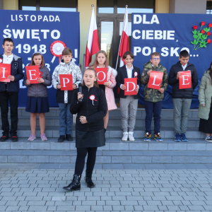 Uczniowie kl. 6 a stoją na schodach przed głównym wejściem do budynku szkolnego. Przed nimi Roksana Romanowska z kl. 4b wykonuje piosenkę tle patriotyczna dekoracja: godło Polski, biało-czerwona flaga oraz laurki przypięte na granatowym tle. Trzymają litery tworzące słowo "Niepodległa" Kliknięcie na zdjęcie spowoduje jego powiększenie do rozmiaru oryginalnego.