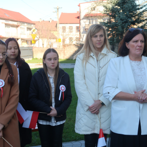 Pani Katarzyna Biedrońska oraz pani Ewa Borowiec ubrane w galowe biało-czarne stroje stoją przed głównym wejściem do budynku szkolnego. Obok uczennice kl. 8 z biało-czerwonymi flagami. Kliknięcie na zdjęcie spowoduje jego powiększenie do rozmiaru oryginalnego.