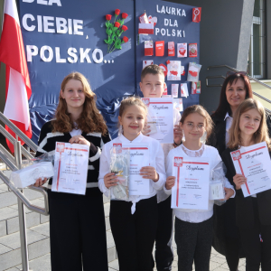 Pani Dyrektor Joanna Ozorka-Krawczyk wraz z laureatami konkursu Laurka dla Niepodległej. W tle okolicznościowa dekoracja z godłem, flagą polski i napisami. Uczestnicy ubrani w galowe stroje trzymają nagrody i dyplomy zdobyte w konkursie. Kliknięcie na zdjęcie spowoduje jego powiększenie do rozmiaru oryginalnego