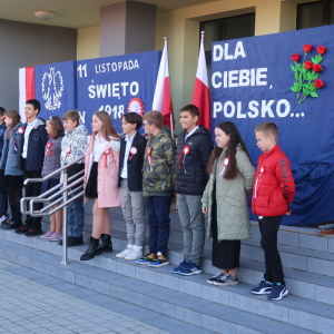 Uczniowie kl. 6 a stoją na schodach przed głównym wejściem do budynku szkolnego. W tle patriotyczna dekoracja: godło Polski, biało-czerwona flaga oraz laurki przypięte na granatowym tle. Kliknięcie na zdjęcie spowoduje jego powiększenie do rozmiaru oryginalnego.