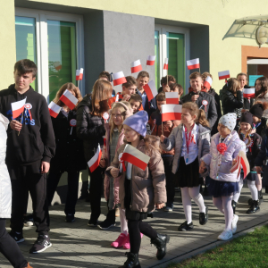 Uczniowie Zespołu Szkół w Szczurowej idą w parach na plac przed głównym wejściem do szkoły. Dyrekcja, nauczyciele i księża ubrani w jesienne ubrania idą wzdłuż budynku szkolnego. Uczestnicy akcji trzymają w rękach biało-czerwone chorągiewki. Kliknięcie na zdjęcie spowoduje jego powiększenie do rozmiaru oryginalnego.