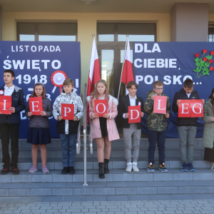 Uczniowie kl. 6 a stoją na schodach przed głównym wejściem do budynku szkolnego. Przed nimi Roksana Romanowska z kl. 4b wykonuje piosenkę tle patriotyczna dekoracja: godło Polski, biało-czerwona flaga oraz laurki przypięte na granatowym tle. Trzymają litery tworzące słowo "Niepodległa" Kliknięcie na zdjęcie spowoduje jego powiększenie do rozmiaru oryginalnego.