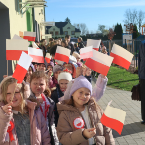 Uczniowie Zespołu Szkół w Szczurowej idą w parach na plac przed głównym wejściem do szkoły. Dyrekcja, nauczyciele i księża ubrani w jesienne ubrania idą wzdłuż budynku szkolnego. Uczestnicy akcji trzymają w rękach biało-czerwone chorągiewki. Kliknięcie na zdjęcie spowoduje jego powiększenie do rozmiaru oryginalnego.