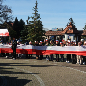 Uczniowie Zespołu Szkół w Szczurowej zebrani na rynku w Szczurowej pod pomnikiem ofiar I i II wojny światowej. Dzieci z kl. 4a i b trzymają przed sobą długą biało-czerwoną flagę. Za nimi pozostali uczestnicy pochodu. Kliknięcie na zdjęcie spowoduje jego powiększenie do rozmiaru oryginalnego.