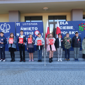 Uczniowie kl. 6 a stoją na schodach przed głównym wejściem do budynku szkolnego. W tle patriotyczna dekoracja: godło Polski, biało-czerwona flaga oraz laurki przypięte na granatowym tle. Kliknięcie na zdjęcie spowoduje jego powiększenie do rozmiaru oryginalnego.