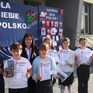 Pani Dyrektor Joanna Ozorka-Krawczyk wraz z laureatami konkursu Laurka dla Niepodległej. W tle okolicznościowa dekoracja z godłem, flagą polski i napisami. Uczestnicy ubrani w galowe stroje trzymają nagrody i dyplomy zdobyte w konkursie. Kliknięcie na zdjęcie spowoduje jego powiększenie do rozmiaru oryginalnego.