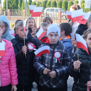 Dyrekcja, księża, nauczyciele i uczniowie Zespołu Szkół w Szczurowej stoją zgromadzeni przed głównym wejściem do szkoły. Uczestnicy wydarzenia ubrani są w ciepłe jesienne ubrania w rękach trzymają biało-czerwone flagi. Kliknięcie na zdjęcie spowoduje jego powiększenie do rozmiaru oryginalnego.