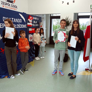 Laureaci Dyktanda niepodległościowego stoją w korytarzu przed głównym wejściem do szkoły. Za nimi tematyczna dekoracja: na granatowym tle biały napis, pod spodem biało-czerwone gołębie obok na granatowym tle biało-czerwone laurki. Młodzież trzyma zdobyte dyplomy, wręcza je pani Wicedyrektor Dorota Gurgul. Kliknięcie na zdjęcie spowoduje jego powiększenie do rozmiaru oryginalnego.