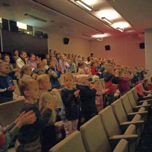 Uczniowie kl. 1-3 w Regionalnym Centrum Kulturalno-Bibliotecznym w Brzesku. Dzieci stoją na sali obok siebie. Słuchają i oglądają projekcję dotyczącą reklamy, a następnie film animowany. Na sali panuje ciemność. Kliknięcie na zdjęcie spowoduje jego powiększenie do rozmiaru oryginalnego.