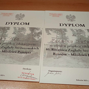 Dyplomy dla uczennic ZS w Szczurowej za udział w projekcie edukacyjnym dotyczącym 80 rocznicy zamordowania Romów w Szczurowej. Leżą na stole obok siebie, dyplomy wydrukowane na białym papierze w tle tematyczne przeźroczyste zdjęcie związane tematycznie z wspomnianym wydarzeniem historycznym. Kliknięcie na zdjęcie spowoduje jego powiększenie do rozmiaru oryginalnego.