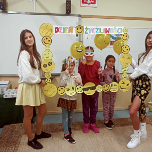 Uczniowie Zespołu Szkół w Szczurowej wraz z uczennicami kl. 8 siedzą wspólnie w sali. Dzieci trzymają żółte balony i są ubrane na żółto. Uczennice kl. 8 trzymają przed nimi ramkę z uśmiechniętymi papierowymi buźkami. Kliknięcie na zdjęcie spowoduje jego powiększenie do rozmiaru oryginalnego.