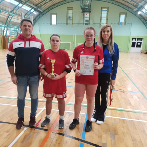 Uczennice Szkoły Branżowej 1 stopnia w Szczurowej stoją w czerwonych strojach sportowych. Uczennice trzymają dyplom  i trofeum zdobyte w zawodach w badmintonie. Obok nauczyciela wychowania fizycznego. Kliknięcie na zdjęcie spowoduje jego powiększenie do rozmiaru oryginalnego.