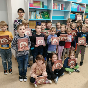 Wychowawczyni kl. 1 a wraz z pierwszoklasistami w szkolnej bibliotece. Pozują na tle regałów. Dzieci ubrane w kolorowe ubrania trzymają przed sobą otrzymane książeczki.