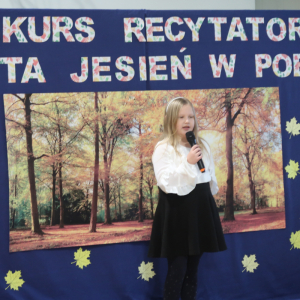 Katarzyna Zabierowska z kl. 2 b recytuje wiersz na konkursie recytatorskim. Ubrana w galowy strój w tle dekoracja z żółtymi liściami, plakatem z drzewami oraz kolorowym napisem. Kliknięcie na zdjęcie spowoduje jego powiększenie do rozmiaru oryginalnego. 