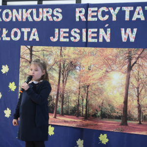 Zuzanna Czaja z kl. 2 b recytuje wiersz na konkursie recytatorskim. Ubrana w galowy strój w tle dekoracja z żółtymi liściami, plakatem z drzewami oraz kolorowym napisem. Kliknięcie na zdjęcie spowoduje jego powiększenie do rozmiaru oryginalnego. 