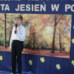 Błażej Szot z kl. 2 b recytuje wiersz na konkursie recytatorskim. Ubrana w galowy strój w tle dekoracja z żółtymi liściami, plakatem z drzewami oraz kolorowym napisem. Kliknięcie na zdjęcie spowoduje jego powiększenie do rozmiaru oryginalnego. 