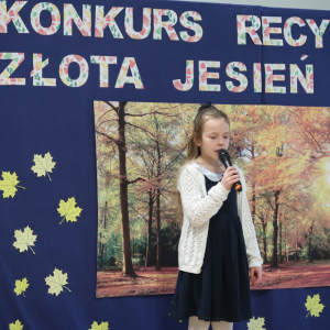 Wiktoria Bogusz z kl. 3 a recytuje wiersz na konkursie recytatorskim. Ubrana w galowy strój w tle dekoracja z żółtymi liściami, plakatem z drzewami oraz kolorowym napisem. Kliknięcie na zdjęcie spowoduje jego powiększenie do rozmiaru oryginalnego. 