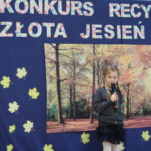 Iga Hanek z kl. 1 b recytuje wiersz na konkursie recytatorskim. Ubrana w galowy strój w tle dekoracja z żółtymi liściami, plakatem z drzewami oraz kolorowym napisem. Kliknięcie na zdjęcie spowoduje jego powiększenie do rozmiaru oryginalnego. 