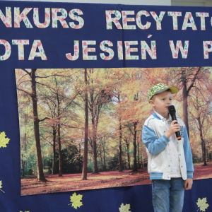 Szymon Budzioch z kl. 1 a recytuje wiersz na konkursie recytatorskim. Ubrany w galowy strój w tle dekoracja z żółtymi liściami, plakatem z drzewami oraz kolorowym napisem. Kliknięcie na zdjęcie spowoduje jego powiększenie do rozmiaru oryginalnego. 