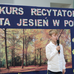 Dominik Gałka z kl. 2 a recytuje wiersz na konkursie recytatorskim. Ubrany w galowy strój w tle dekoracja z żółtymi liściami, plakatem z drzewami oraz kolorowym napisem. Kliknięcie na zdjęcie spowoduje jego powiększenie do rozmiaru oryginalnego. 