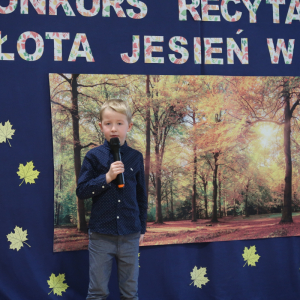 Maksymilian Gawełczyk z kl. 1 a recytuje wiersz na konkursie recytatorskim. Ubrany w galowy strój w tle dekoracja z żółtymi liściami, plakatem z drzewami oraz kolorowym napisem. Kliknięcie na zdjęcie spowoduje jego powiększenie do rozmiaru oryginalnego. 