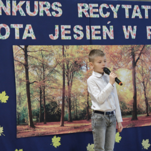 Jakub Maj z kl. 2 a recytuje wiersz na konkursie recytatorskim. Ubrany w galowy strój w tle dekoracja z żółtymi liściami, plakatem z drzewami oraz kolorowym napisem. Kliknięcie na zdjęcie spowoduje jego powiększenie do rozmiaru oryginalnego. 