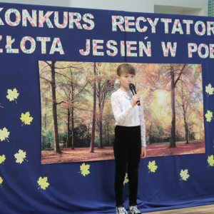 Maja Kozak z kl. 3 b recytuje wiersz na konkursie recytatorskim. Ubrana w galowy strój w tle dekoracja z żółtymi liściami, plakatem z drzewami oraz kolorowym napisem. Kliknięcie na zdjęcie spowoduje jego powiększenie do rozmiaru oryginalnego. 