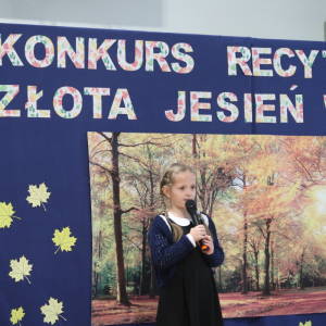 Iwona Rosa z kl. 1 b recytuje wiersz na konkursie recytatorskim. Ubrana w galowy strój w tle dekoracja z żółtymi liściami, plakatem z drzewami oraz kolorowym napisem. Kliknięcie na zdjęcie spowoduje jego powiększenie do rozmiaru oryginalnego. 