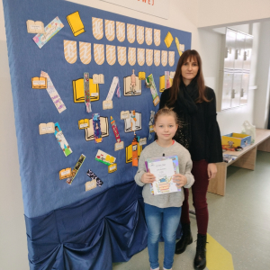 Klara Markowicz laureatka Szkolnego Konkursu Czytelniczego "Zakładka do książki" w towarzystwie pani Wicedyrektor Doroty Gurgul. W tle granatowa dekoracja z kolorowymi zakładkami oraz biało-pomarańczowym napisem i elementami dekoracyjnymi w kształcie książek w kolorach żółtych i pomarańczowych. Kliknięcie na zdjęcie spowoduje jego powiększenie do rozmiaru oryginalnego.