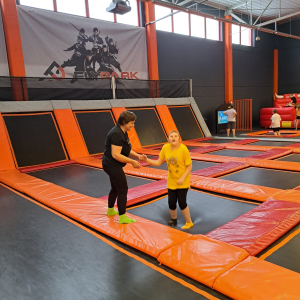 Uczennica z pomocą nauczyciela bawi się na trampolinach. Kliknięcie na zdjęcie spowoduje jego powiększenie do rozmiaru oryginalnego.