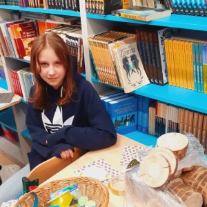 Uczennica kl. 7b siedzi przy stole w szkolnej bibliotece. Przygotowuje kartki z życzeniami do  bombek dla starszych i samotnych. Ozdoby świąteczne wykonane z cekinów i innych ozdobnych materiałów umieszczono na drewnianych spodkach i ozdobiono dekoracjami. Kliknięcie na zdjęcie spowoduje jego powiększenie do rozmiaru oryginalnego.