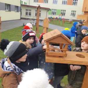 Uczniowie z kl. 1 b stoją przy drewnianych karmnikach. Ubrani są w ciepłe ubrania i czapki. Jeden z chłopców wkłada pokarm do karmnika. Kliknięcie na zdjęcie spowoduje jego powiększenie do rozmiaru oryginalnego.