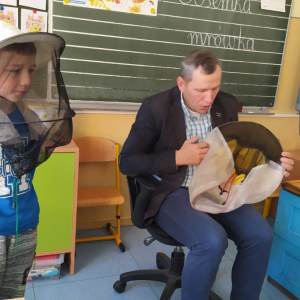 Pan Józef przybyło pokazuje kapelusz ochronny pszczelarza. Obok niebieski ul przyniesiony do klasy oraz chłopiec z założonym na głowę kapeluszem. Kliknięcie na zdjęcie spowoduje jego powiększenie do rozmiaru oryginalnego.