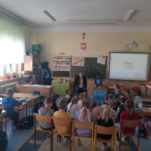 Pan Józef Przybyło pokazuje dzieciom niebiesko-brązowy ul. Opowiada o życiu pszczół. Dzieci siedzą zgromadzone przed nim na dywanie. W tle tablica zielona z białymi liniami oraz biało-szara tablica multimedialna. Kliknięcie na zdjęcie spowoduje jego powiększenie do rozmiaru oryginalnego.