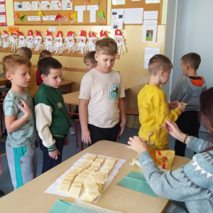 Wychowawczyni kl. 1 b siedzi przy stoliku. Smaruje miodem wafelki, a następnie podaje je dzieciom, które czekają w kolejce. Kliknięcie na zdjęcie spowoduje jego powiększenie do rozmiaru oryginalnego.