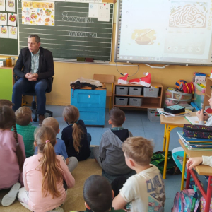 Pan Józef Przybyło siedzi na krześle na środku klasy 1 b. Przed nim na dywanie zgromadzeni są uczniowie, którzy słuchają jego opowiadania o pszczołach. W tle tablica z liniami i tablica multimedialna.  Kliknięcie na zdjęcie spowoduje jego powiększenie do rozmiaru oryginalnego.