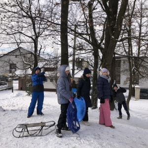 Młodzież stoi na górce. Na ziemi obok nich sanki. Niektórzy trzymają jabłuszka i inny sprzęt do zjeżdżania po śniegu. W tle drzewa. Kliknięcie na zdjęcie spowoduje jego powiększenie do rozmiaru oryginalnego.