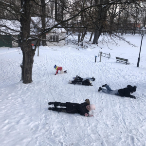 Chłopcy i dziewczynki turlają się z górki wokół własnej osi po śniegu. Obok drzewa. Dzieci ubrane są w zimowe ubrania i buty. Kliknięcie na zdjęcie spowoduje jego powiększenie do rozmiaru oryginalnego.